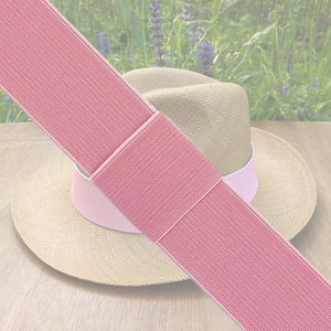 May include: A straw hat with a pink band and a pink elastic band. The hat has a wide brim and a light-coloured crown. The elastic band is a solid pink colour with a textured pattern.