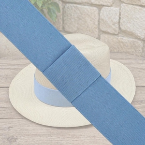 May include: A light blue ribbon is draped over a straw hat with a light blue band. The ribbon is made of a textured fabric and is approximately 2 inches wide. The hat is a classic fedora style.