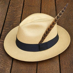 May include: A light tan Panama hat with a black band and a decorative feather. The hat has a wide brim and a classic fedora shape. The feather is brown with dark brown markings.