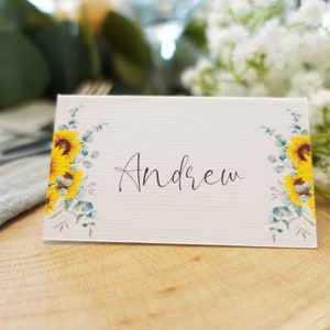 Puede incluir: Tarjeta de lugar blanca con un dise&ntilde;o de girasol y eucalipto. El nombre "Andrew" est&aacute; escrito en caligraf&iacute;a negra.