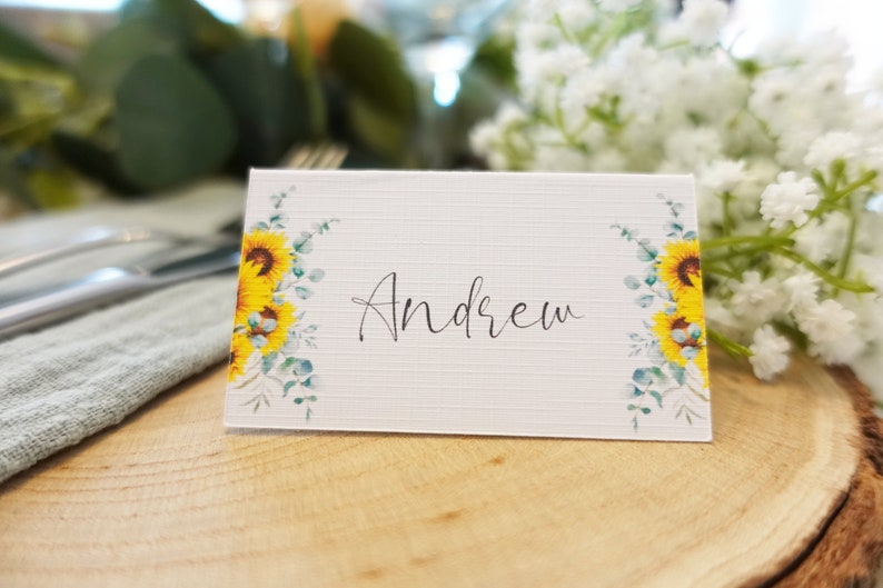 Puede incluir: Tarjeta de lugar blanca con un dise&ntilde;o de girasol y eucalipto. El nombre "Andrew" est&aacute; escrito en caligraf&iacute;a negra.