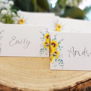 Puede incluir: Dos tarjetas de lugar blancas con dise&ntilde;os de girasol y eucalipto. Las tarjetas tienen los nombres "Emily" y "Andrew" escritos en caligraf&iacute;a negra.