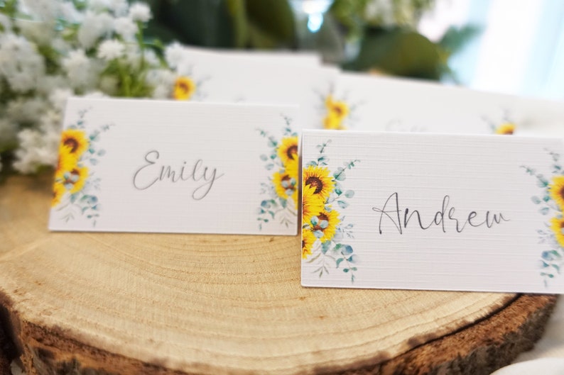Puede incluir: Dos tarjetas de lugar blancas con dise&ntilde;os de girasol y eucalipto. Las tarjetas tienen los nombres "Emily" y "Andrew" escritos en caligraf&iacute;a negra.