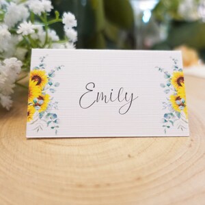 Puede incluir: Tarjeta de lugar blanca con un dise&ntilde;o de girasol y eucalipto. El nombre "Emily" est&aacute; escrito en cursiva negra.