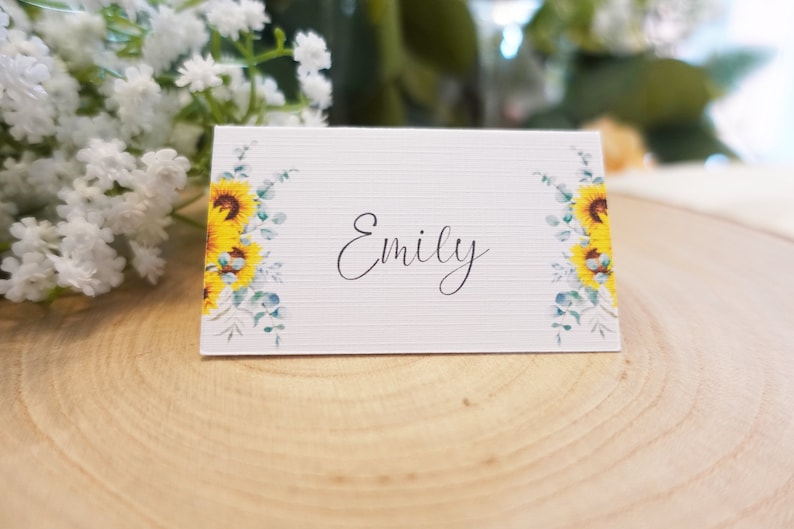Puede incluir: Tarjeta de lugar blanca con un dise&ntilde;o de girasol y eucalipto. El nombre "Emily" est&aacute; escrito en cursiva negra.