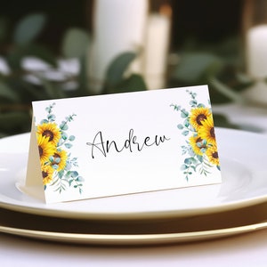 Puede incluir: Una tarjeta de lugar blanca con un dise&ntilde;o de girasol y eucalipto. El nombre "Andrew" est&aacute; escrito en cursiva negra.
