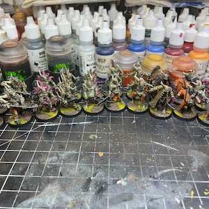 May include: A collection of 18 miniature figures painted in various shades of green, brown, and orange. The figures are arranged in a line on a black and white grid surface. The figures are all wearing armor and carrying weapons.