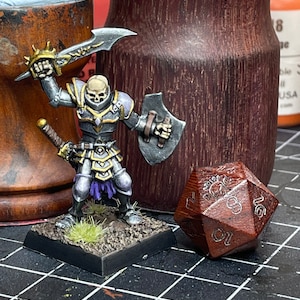 May include: A painted miniature of a skeletal warrior wearing silver armor and holding a sword and shield. The miniature is standing on a black base with green grass. A brown wooden 20-sided die is in the foreground.