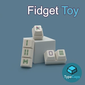 Op de afbeelding: Witte Fidget Toy-toetsenbordtoetsen met groene en paarse symbolen en cijfers. De tekst "Fidget Toy" staat bovenaan de afbeelding. Het TypeCaps-logo staat rechtsonder.