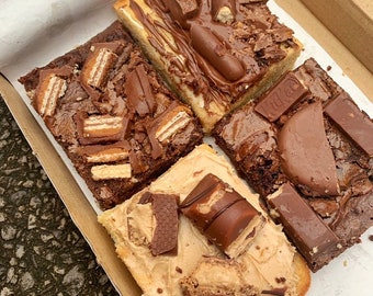 May include: Four different brownie squares with various chocolate toppings, including chocolate bars, chocolate chips, and chocolate sauce. The brownies are arranged in a cardboard box lined with white paper.