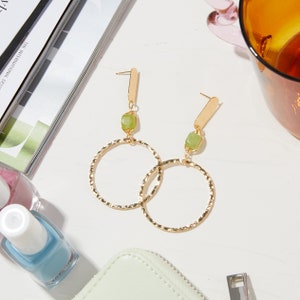 May include: Gold hoop earrings with a textured finish and a green stone dangle. The earrings have a simple, modern design.