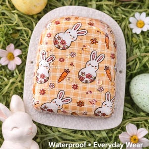 May include: A decorative, patterned item featuring a repeating design of white bunnies with floral accents and carrots on a yellow plaid background. The item is surrounded by Easter eggs and flowers. The text "Waterproof • Everyday Wear" is visible.