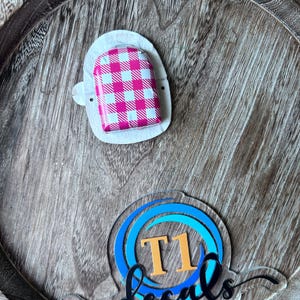 May include: A pink and white gingham patterned object on a white backing. The object is on a wooden tray. A clear acrylic decal with the text "T1 decals" in black script and orange and blue circles is also on the tray.