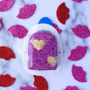 Può includere: Un cioccolatino a forma di cuore viola avvolto in foglia d'oro, posto su un cerotto adesivo bianco a forma di cuore. Il cerotto è circondato da cuori di glitter rosa e rossi.