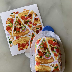 May include: A white card and a medical device with a pizza slice pattern. The pizza design includes pepperoni, mushrooms, green peppers, and red onions. The medical device has a blue plastic component and is wrapped in white fabric.