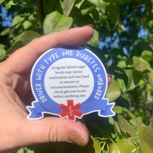 May include: Blue and white sticker with the text "DRIVER WITH TYPE ONE DIABETES ONBOARD". The sticker has a red medical symbol with a snake wrapped around a staff. The text reads: "Irregular blood sugar levels may mimic inebriation and may lead to seizure or unconsciousness. Please check glucose levels before anything else."