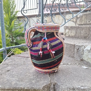 Pode incluir: Um jarro decorativo coberto por um tecido com riscas diagonais em tons de roxo, verde, vermelho e preto. O jarro tem um rebordo de corda grossa, uma pega resistente e duas borlas pendentes com pequenas contas azuis e fibras escuras.