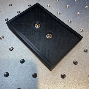 Op de afbeelding: Een zwart rechthoekig object met twee metalen inzetstukken, mogelijk een montageplaat of basis. Het object heeft een verhoogde rand en ligt op een zilveren oppervlak met talrijke schroefdraadgaten, wat wijst op een precisieproductie- of assemblageomgeving.