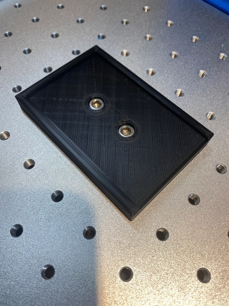 Op de afbeelding: Een zwart rechthoekig object met twee metalen inzetstukken, mogelijk een montageplaat of basis. Het object heeft een verhoogde rand en ligt op een zilveren oppervlak met talrijke schroefdraadgaten, wat wijst op een precisieproductie- of assemblageomgeving.
