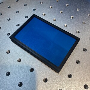Op de afbeelding: Een rechthoekig object met een zwart frame en een levendig blauw interieur. Het object is geplaatst op een metalen oppervlak met gelijkmatig verdeelde gaten. Het frame lijkt van een mat materiaal te zijn.