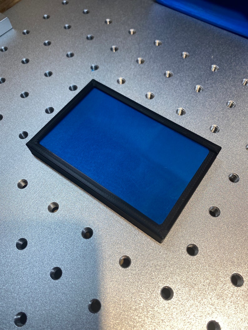 Op de afbeelding: Een rechthoekig object met een zwart frame en een levendig blauw interieur. Het object is geplaatst op een metalen oppervlak met gelijkmatig verdeelde gaten. Het frame lijkt van een mat materiaal te zijn.