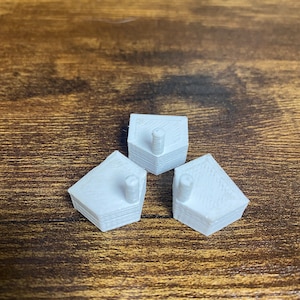 Puede incluir: Tres pequeños objetos blancos con forma de casa, impresos en 3D, con una superficie texturizada. Cada uno tiene un pequeño poste cilíndrico que se extiende desde la parte superior. Los objetos están dispuestos sobre una superficie de madera.