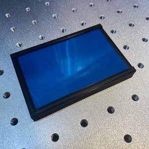 Op de afbeelding: Een rechthoekig object met een zwart frame en een levendig blauw interieur. Het frame lijkt van een mat materiaal te zijn gemaakt. Het object rust op een metalen oppervlak met gelijkmatig verdeelde gaten.