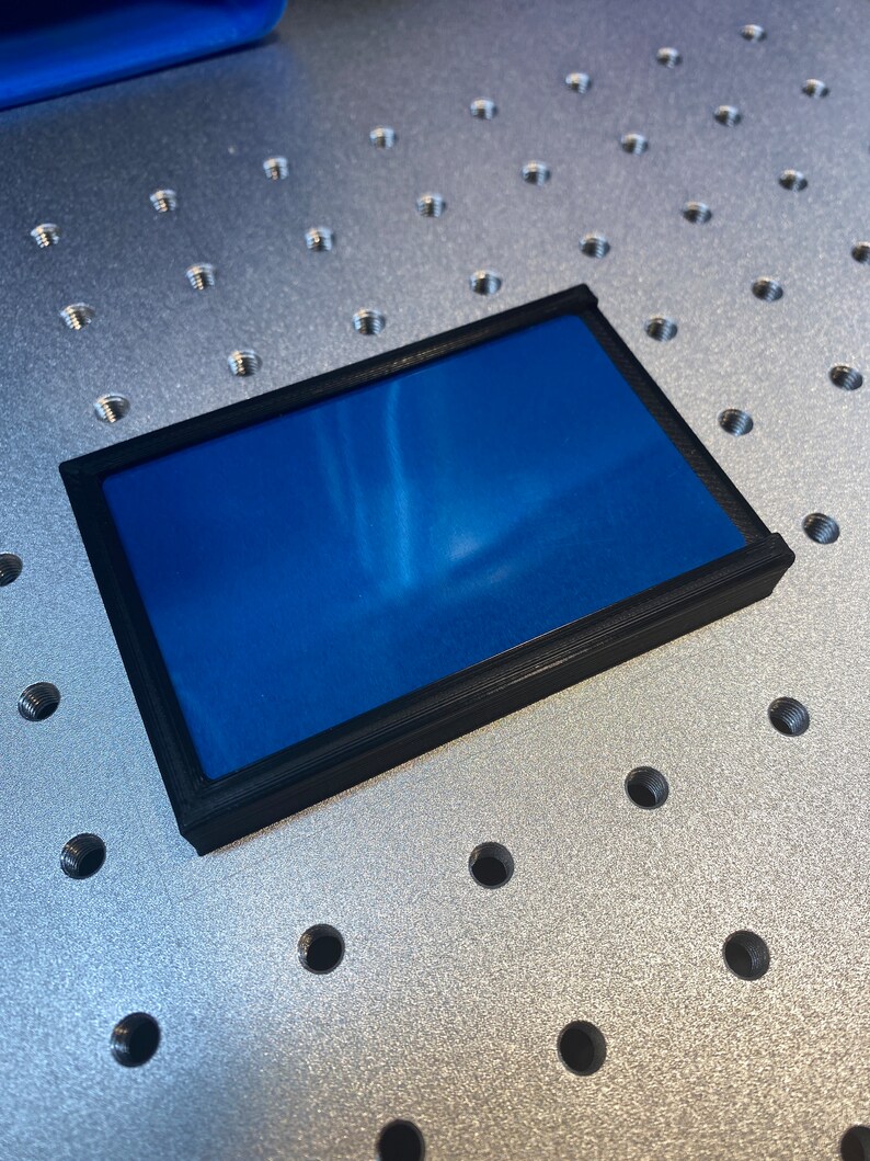 Op de afbeelding: Een rechthoekig object met een zwart frame en een levendig blauw interieur. Het frame lijkt van een mat materiaal te zijn gemaakt. Het object rust op een metalen oppervlak met gelijkmatig verdeelde gaten.