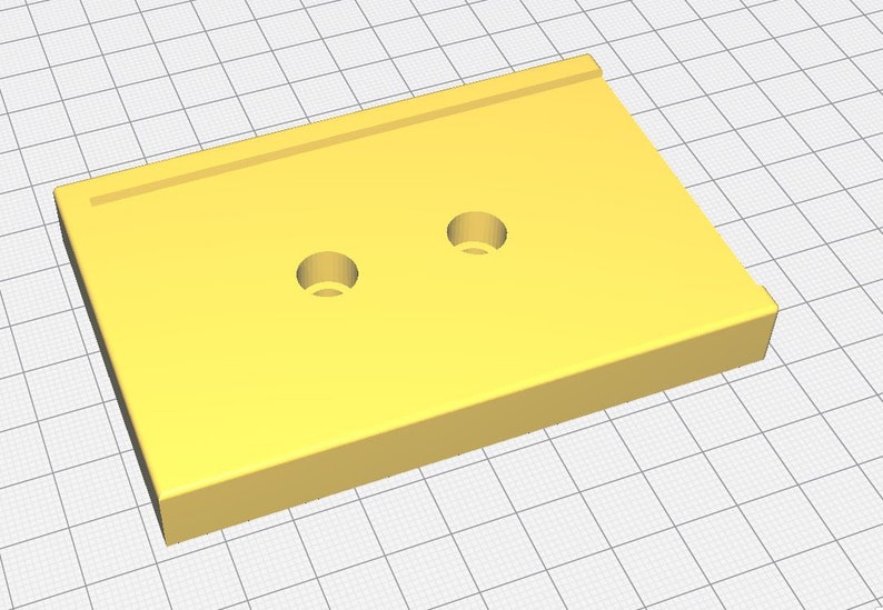 Op de afbeelding: Een effen geel rechthoekig object met twee cirkelvormige gaten. De bovenkant heeft een verhoogde rand langs &eacute;&eacute;n kant. Het object staat tegen een rasterachtergrond, wat wijst op een 3D-model of ontwerpelement.