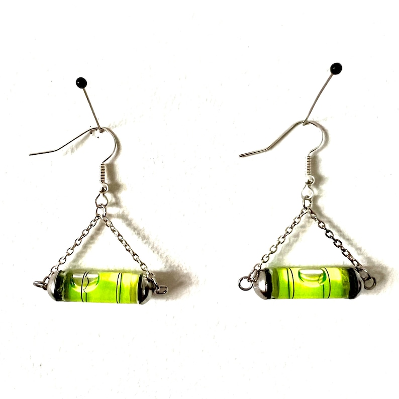 May include: A pair of silver earrings with a green level bubble attached to each earring. The earrings are hanging from silver hooks.
