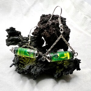 May include: A pair of silver earrings with green level bubbles hanging from a silver chain. The earrings are on a dark gray rock.
