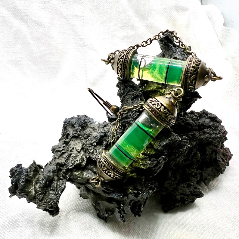 May include: A pair of green liquid filled earrings with ornate gold-toned metal caps and chains. The earrings are displayed on a dark gray textured surface.