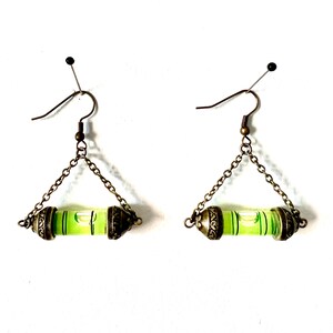 May include: A pair of bronze earrings with a green level bubble hanging from a chain. The level bubble is encased in a clear glass tube with a bronze cap on each end.