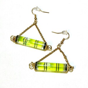 May include: A pair of gold earrings with a level bubble charm. The level bubbles are green and have a black line in the center. The earrings are hanging from a gold chain.