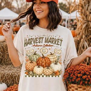 May include: An off-white short-sleeve t-shirt featuring a graphic print of various pumpkins and the text Harvest Market Autumn Collection. The design includes pumpkins with different patterns and colours, surrounded by autumn foliage and a chequered border.