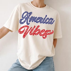 Pode incluir: Camiseta creme com a frase "America Vibes" em azul e vermelho. O texto azul é preenchido com estrelas brancas, e o texto vermelho é preenchido com pontos brancos. A camisa é combinada com jeans azul claro e tênis brancos com listras pretas.