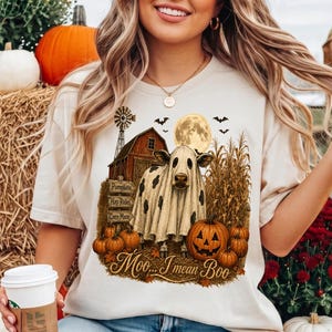 Pode incluir: Uma t-shirt cor de creme com um gráfico de uma vaca vestida de fantasma em frente a um celeiro, um milharal e abóboras. O texto diz Moo... I mean Boo. A t-shirt é combinada com calças de ganga azul claro com efeito desgastado.