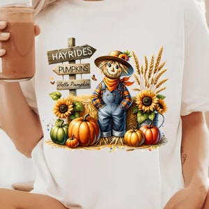 Pode incluir: T-shirt branco com um gráfico colorido com tema de outono. O design inclui um espantalho, abóboras, girassóis e uma placa que diz "Hayrides, Pumpkins, Hello Pumpkin". A camisa é um estilo casual.