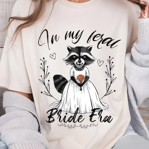 May include: A white t-shirt with a black and white illustration of a raccoon wearing a wedding dress and holding a bouquet of flowers. The text "In my feral bride era" is printed on the shirt.