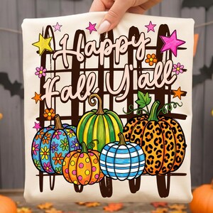 Pode incluir: Uma t-shirt de cor creme com um gráfico de cinco abóboras com vários padrões, incluindo flores, riscas e estampado de leopardo. O texto "Happy Fall Y'all" está impresso numa fonte cursiva sobre as abóboras, acentuado por estrelas coloridas.