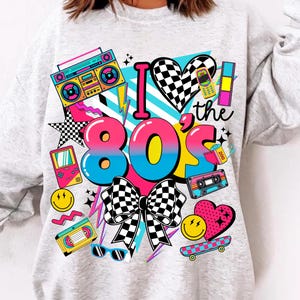 May include: A light gray sweatshirt with a colorful 80s-themed graphic. The design includes the text "I ❤️ the 80s" with a checkered heart, bow, and various retro icons like a boombox, cassette tape, and a handheld game console.