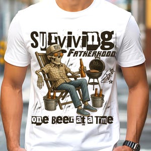 Pode incluir: Camiseta branca com um gráfico de um esqueleto usando óculos de sol e boné de beisebol, sentado em uma cadeira com uma garrafa de cerveja. O texto diz "SURVIVING FATHERHOOD one BEER at a TIME". Uma churrasqueira e baldes de cerveja também estão na imagem.