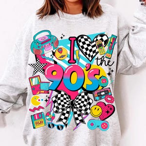 May include: A light gray sweatshirt with a colorful 90s-themed graphic. The design includes the text "I love the 90's" with a checkered heart and bow, along with retro icons like a boombox, cassette tape, and video game controller.