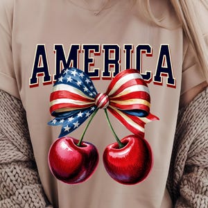 Pode incluir: Uma camiseta bege com a palavra "AMERICA" impressa em letras azuis. Um laço vermelho, branco e azul com estrelas está centralizado na camiseta. Duas cerejas vermelhas com hastes verdes estão abaixo do laço.
