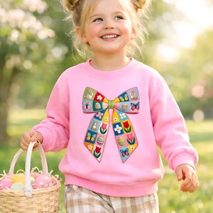Pode incluir: Um suéter rosa infantil com um grande design de laço colorido com motivos de Páscoa. A criança está segurando uma cesta de vime cheia de ovos de Páscoa em tons pastel. O fundo é uma cena externa desfocada.