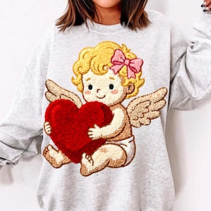 Pode incluir: Camisola cinza claro com um desenho bordado de um cupido de desenho animado segurando um grande coração vermelho. O cupido tem cabelo loiro encaracolado, um laço rosa e asas. O desenho está na frente da camisola.