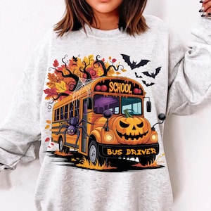 May include: A light gray sweatshirt featuring a Halloween-themed design. The graphic depicts an orange school bus with a jack-o'-lantern face, a spider, and the words "SCHOOL BUS DRIVER". The design includes bats and autumn leaves.