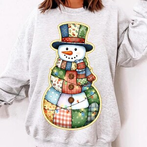 Pode incluir: Uma camisola cinza claro com um design colorido de boneco de neve. O boneco de neve tem um corpo de retalhos, um chapéu de coco preto e um cachecol estampado. O design inclui botões e luzes de corda.