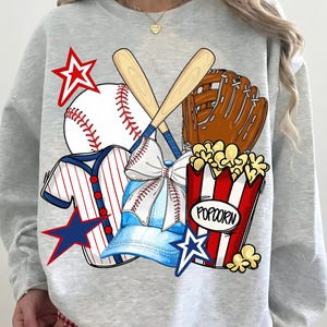 May include: A light grey sweatshirt with a baseball design. The graphic includes a baseball, bats, glove, popcorn, a baseball cap, and star accents in red, white, and blue. The word "Popcorn" is printed on the popcorn container.