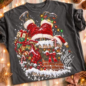 Pode incluir: Uma t-shirt cinzento-carvão com um gráfico do Pai Natal preso de cabeça para baixo numa chaminé de tijolo. O design inclui pernas às riscas vermelhas e brancas, botas pretas, presentes, neve, luzes de Natal e o texto Well, Shit...